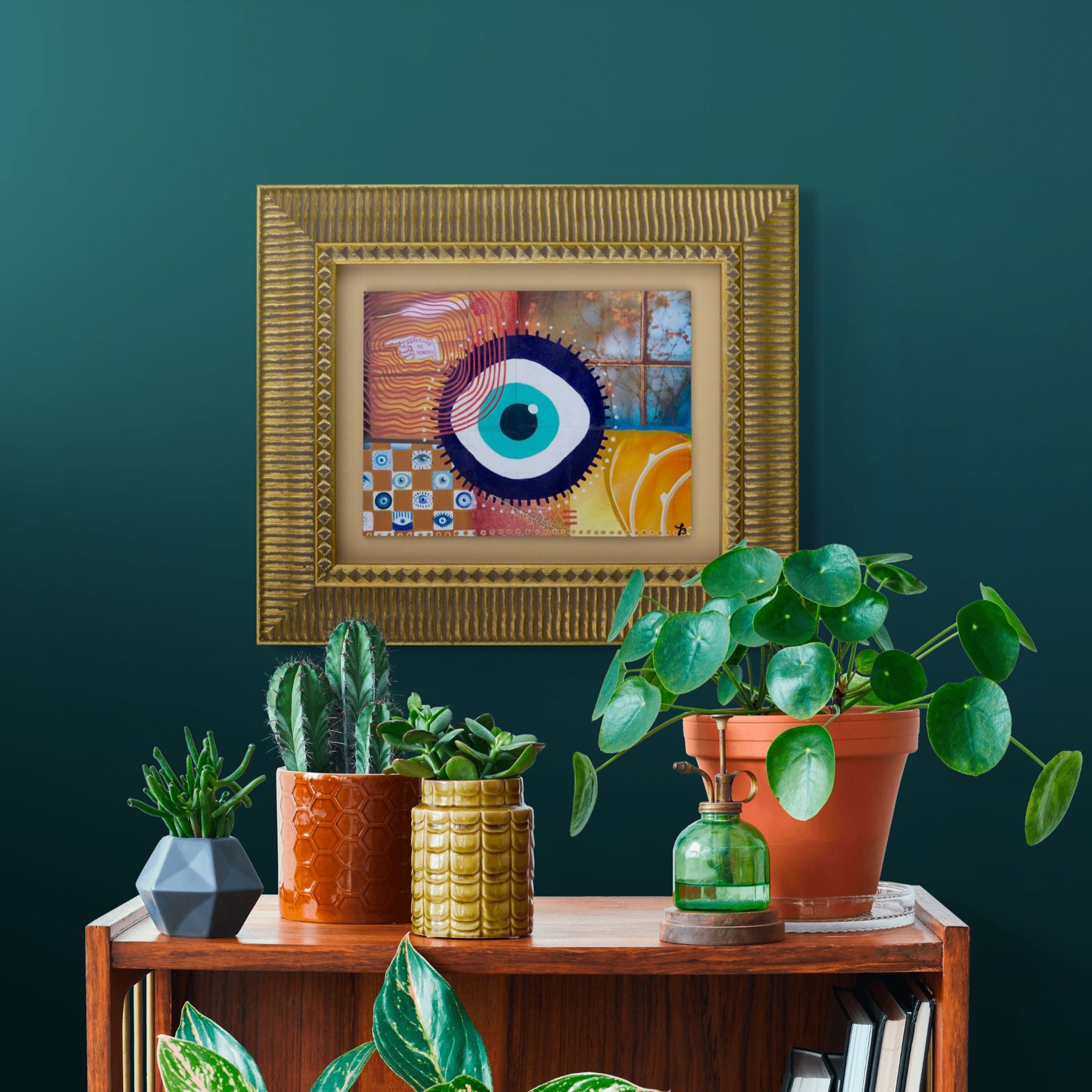 Decorative setup with framed original mixed media artwork of an evil eye and potted plants against a dark green wall.