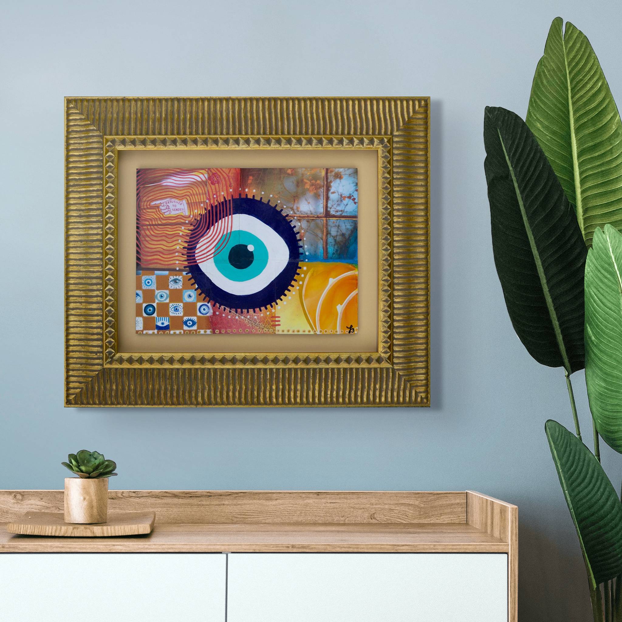 Mixed media collage original wall art with a colorful evil eye design in a gold frame on a light blue wall.