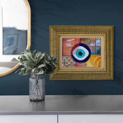 Mixed media collage art with an evil eye framed on a wall with a vase of plants in the foreground.