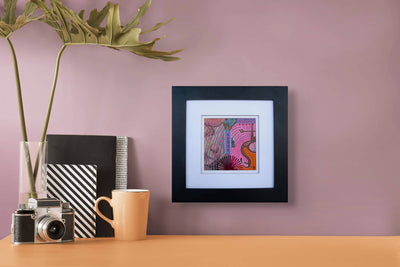 Framed collage original artwork on a wall with a table displaying a camera, mug, and books against a pink background.