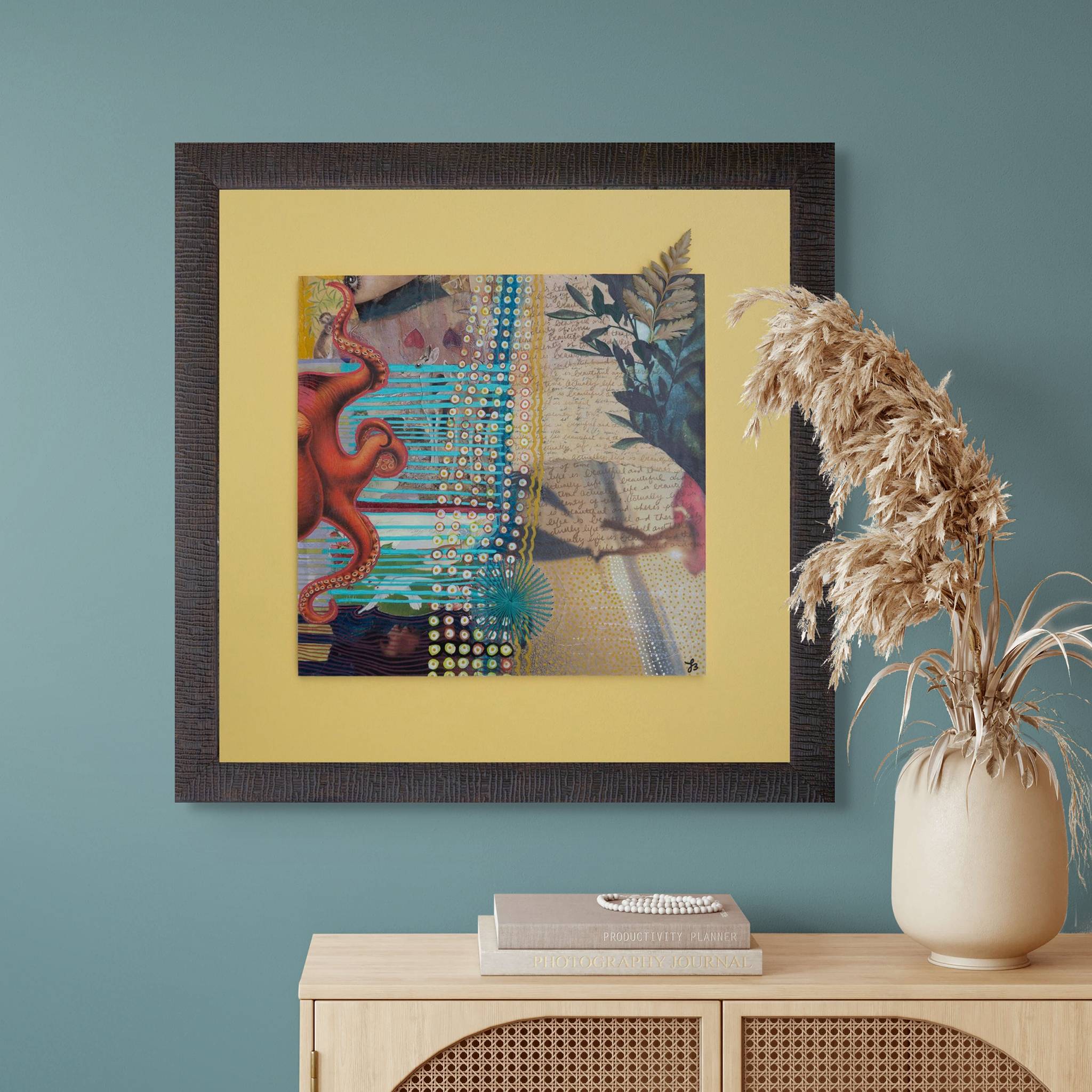 Framed abstract orginal collage art piece on a teal wall with a decorative vase and books on a wooden cabinet.