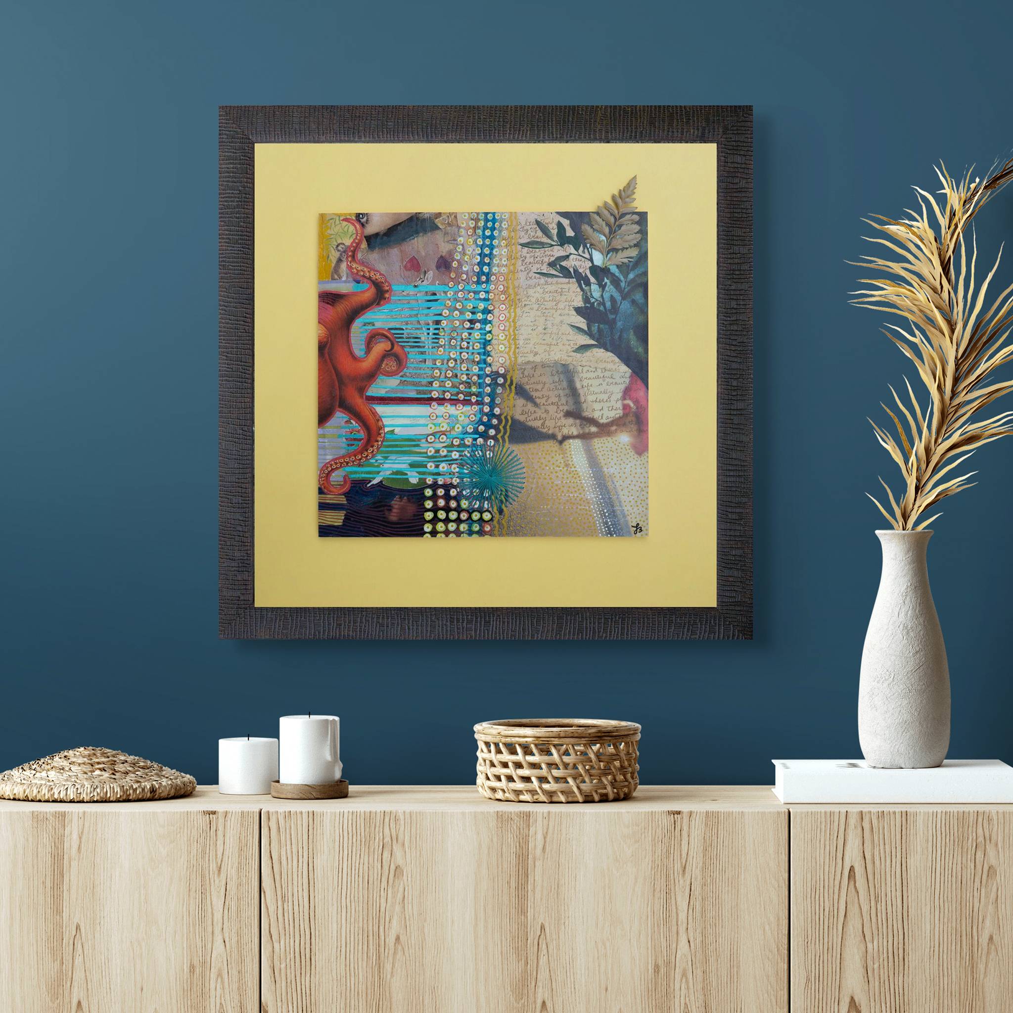 Framed abstract mixed media collage art piece on a blue wall with decorative items on a wooden shelf.